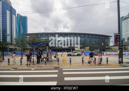 Fußgänger warten neben einem breiten Zebrastreifen vor dem Busan-Bahnhof, modernen Hochhäusern und einer bewölkten städtischen Skyline. Stockfoto