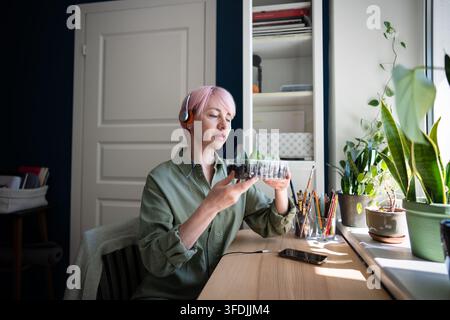 Frau mit Kopfhörern überprüft Zamioculcas-Ableger neben Zimmerpflanzen in einem sonnigen Wohnraum. Stockfoto