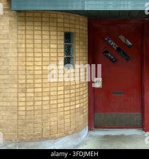 Eine rote Tür neben einer geschwungenen Wand aus quadratischen hellbraunen Fliesen bildet einen markanten streamline-modernen architektonischen Eingang. Stockfoto