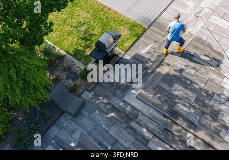 Von oben gesehen sprüht ein Arbeiter in Stiefeln eine gepflasterte Terrasse neben einem Rasen. Schläuche ziehen sich über die Oberfläche, und ein Grill steht nahe der Gartenkante. Stockfoto