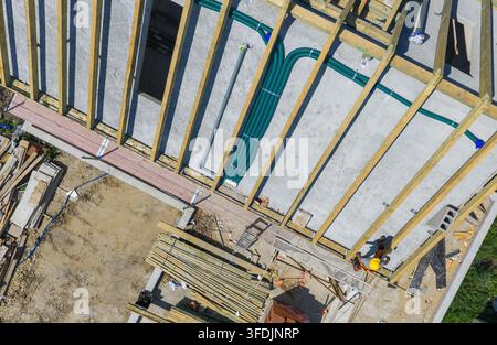 Eine Ansicht von oben zeigt parallele Dachbalken aus Holz, freiliegende Rohre und Gerüstplattformen. Ein Arbeiter und gestapelte Baumaterialien erscheinen entlang der Kante Stockfoto