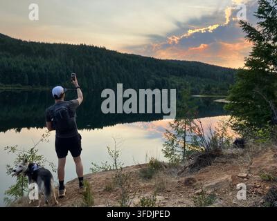 Ein Wanderer fotografiert einen leuchtenden Sonnenuntergang, der sich auf dem ruhigen Allen Lake spiegelt, mit seinem Husky neben ihm in den bewaldeten Bergen in der Nähe von Cumberland, BC Stockfoto