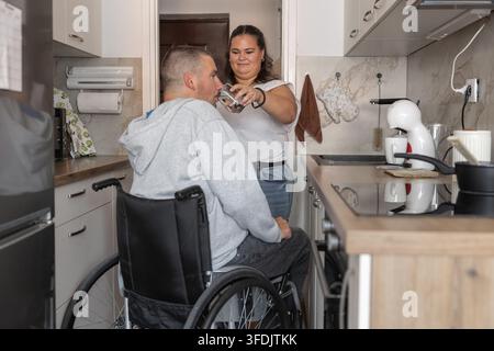 Persönliche Assistentin hilft einem Mann mit einer Behinderung, während einer täglichen Routine in einer häuslichen Umgebung, Wasser zu trinken. Stockfoto