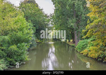 Grüner Kanal mit üppigen grünen Bäumen und zarter Fontänenfontäne, ruhige Naturszene an einem bewölkten Tag. Stadt Brügge in Belgien, Europa. Stockfoto