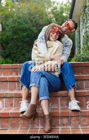 Zwei Frauen umarmen und lachen auf roten Ziegelstufen und tragen lustige Brillen vor dem Haus Stockfoto