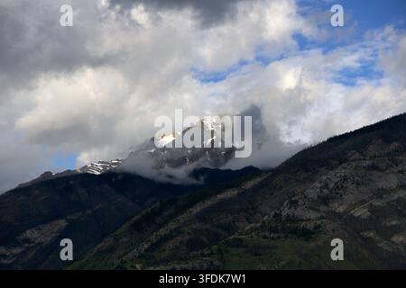 Ein hoher schneebedeckter Alpengipfel, umgeben von flauschigen wirbelnden weißen Wolken unter einem Flecken blauen Himmels und dramatisch dunklen, bewaldeten Berghängen Stockfoto