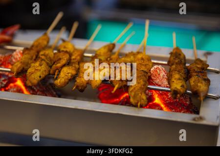 Reihen von gewürztem Hühnersatay-Spießen, die über glühenden roten Holzkohle-Glut in einer Metallrinne an einem Nachtmarktstand gegrillt werden. Stockfoto