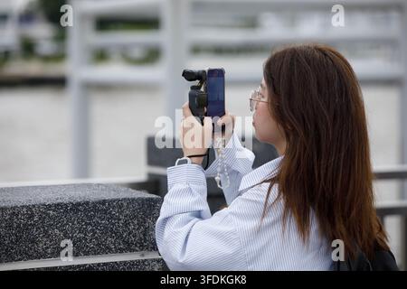 BANGKOK, THAILAND - 15. AUGUST 2026: Frau verwendet Smartphone und Vlog-Kamera, um Fotos aufzunehmen, Seitenansicht, Tha Tien. Stockfoto
