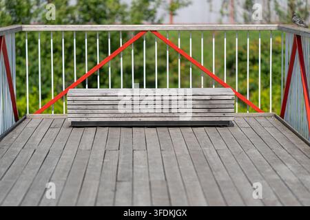 Eine verwitterte Holzbank steht einladend auf einem glatten Deck, das von roten und weißen Geländern eingerahmt ist. Die umgebende Grünfläche verleiht einen Hauch von der Schönheit der Natur c Stockfoto