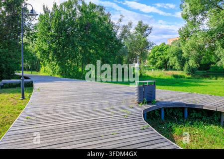 Gebogener Holzsteg windet sich durch einen üppigen grünen Park. Heller blauer Himmel mit weißen Wolken darüber Stockfoto