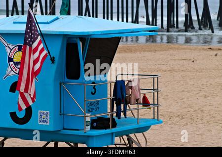 Heller blauer Rettungsturm mit einer amerikanischen Flagge steht auf einem Sandstrand neben Pierpfählen und Ozeanwellen in Pismo Beach, Kalifornien. Stockfoto