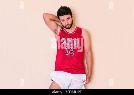 Athletischer junger Mann trägt ein rotes Team-Tanktop und eine schwarze Kappe, mit einem erhobenen Arm in entspannter Pose, der einen starken Bizeps zeigt. Sportfan trägt Stockfoto