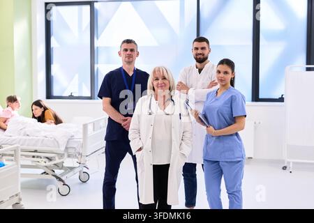Medizinisches Team steht zusammen in einem hellen Krankenzimmer, während im Hintergrund Patienten während eines geschäftigen Tages versorgt werden Stockfoto