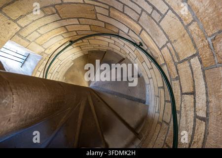 Eine Hochwinkel-Innenansicht, die eine schmale, gewundene mittelalterliche Steinspiraltreppe mit einem geschwungenen Metallgeländer hinunterblickt. Stockfoto
