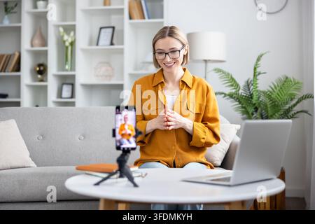Frau, die einen Live-Videoanruf überträgt oder ein Vlog von ihrem Homeoffice aus erstellt, mit einem Smartphone auf einem Stativ und einem Laptop, um sich mit einem Publikum zu verbinden und Inhalte online zu teilen Stockfoto
