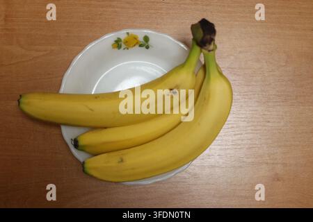 Drei reife Bananen liegen auf einem weißen Teller, der mit gelben Blumen verziert ist. Der Teller ruht auf einem Holztisch, der eine schlichte Kücheneinrichtung zeigt. Stockfoto