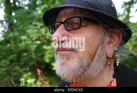 Mann im Profil, in seinen Sechzigern, trägt einen strukturierten schwarzen Hut und eine Brille. Er hat einen gepflegten grauen Bart. Mehrere Ohrringe, darunter ein Anker-Ankh-Zeichen Stockfoto