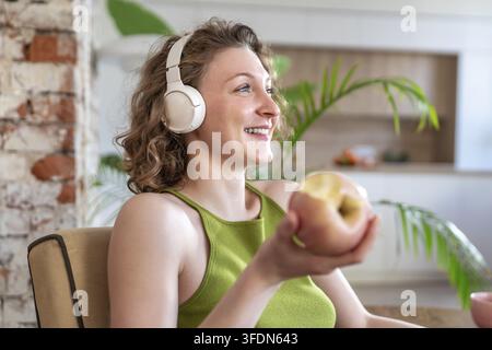 Sorglose, lächelnde junge Frau genießt einen gesunden Snack und hört ihre Lieblingsmusik zu Hause. Entspannte Frau in kabellosen Kopfhörern hält einen frischen Apfel, fühlt Stockfoto