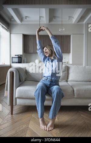 Zufriedene, zufriedene Frau lächelt mit geschlossenen Augen, streckt sich nach der Ruhe und sitzt auf der Couch. Gelassene, entspannte Frau mittleren Alters genießt einen achtsamen Moment zu Hause Stockfoto