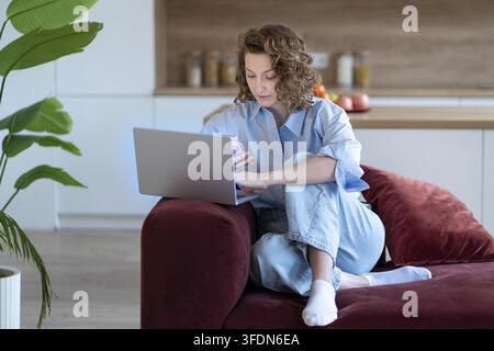 Moderne Freiberuflerfrau, die von zu Hause aus remote arbeitet, Multitasking mit Laptop und Smartphone, während sie auf einem gemütlichen Sofa sitzt. Konzentrierte tech-affine Frau Stockfoto