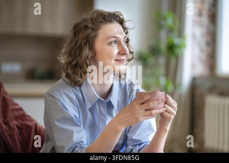 Träumende junge Frau trinkt Kaffee, genießt einen gemütlichen Moment zu Hause. Lächelnde Frau fühlt sich entspannt, macht eine Pause von den täglichen Aufgaben mit einer Tasse Tee. Achtsamkeit Stockfoto