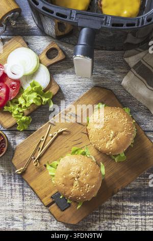 Zwei herzhafte köstliche hausgemachte Cheeseburger in einem modernen Luftfritteuse-Küchengerät auf Holzunterlage. Lifestyle-Trend des neuen Kochens. Top-Down-Flachlayout Stockfoto