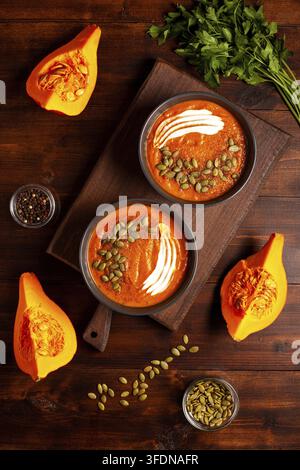 Kürbissuppe mit Samen, Sauerrahm, Gewürzen in einer dunklen Schüssel, Kräuter und geschnittene Kürbisse auf einem Holztisch, von oben aufgenommen. Das Konzept von gesundem Gemüse Stockfoto