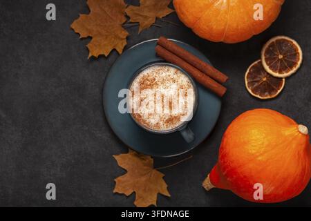 Flat Lay einer gemütlichen Herbstgetränkeszene mit einem Pumpkin Spice Latte in einer dunklen Keramiktasse, Zimtstangen, Kürbissen, getrockneten Orangen und Ahornblättern auf Stockfoto