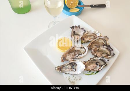 Frische Austern auf einem weißen Teller angeordnet mit Zitronenscheiben, begleitet von einem Glas Weißwein in einer hellen Essumgebung Stockfoto