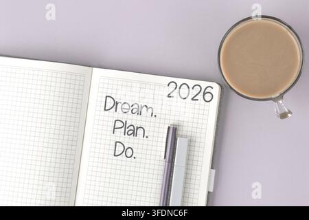 Offenes Notizbuch mit dem motivierenden Satz Träume. Plane. Handle. Jahr 2026 auf einer Rasterseite, neben einer Tasse Kaffee und zwei Stiften auf pastelllavendelfarbenem Untergrund Stockfoto