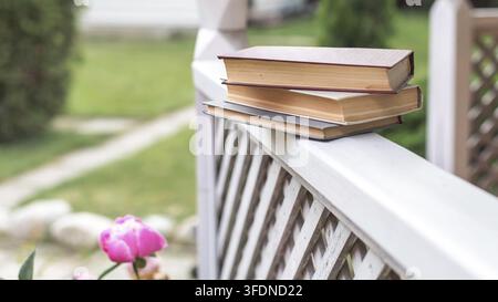 Stapel alter Hardcover-Bücher, die auf einem Holzgeländer in einem Garten mit sanft verschwommener Vegetation und blühender pinker Blume platziert sind, natürliches Licht, friedliches Lesen Stockfoto