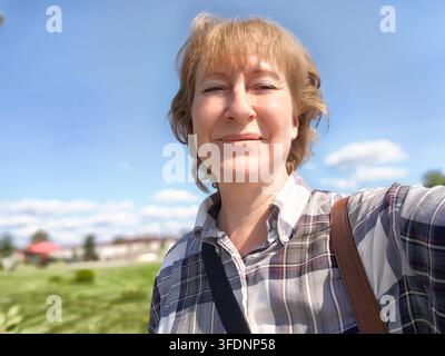 Eine Frau lächelt, während sie ein Selfie in einer hellen Außeneinrichtung mit einer grünen Landschaft und einem klaren blauen Himmel macht. Stockfoto