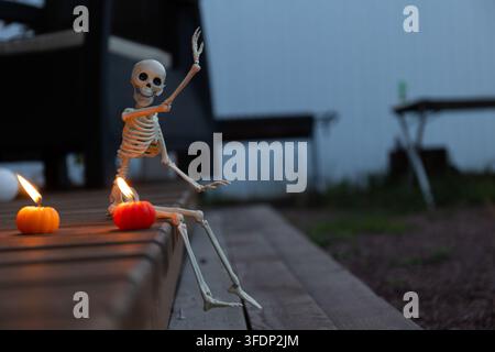 Ein Plastik-Skelett sitzt neben flackernden Kerzen und Kürbissen auf einer Holzbank, während die blaue Dämmerung hereinbricht, und schafft so eine mystische, von Kerzenlicht erhellte Halloween-Szene Stockfoto