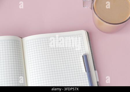 Ein offenes Gitter-Notizbuch mit einem Stift und einer Tasse Kaffee, die auf einem rosa Hintergrund platziert sind. Das minimalistische Arbeitsplatz-Setup repräsentiert Fokus und Produktivität. Co Stockfoto