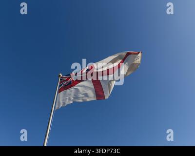 Royal Navy White Ensign-Flagge, die von einem Flaggenmast vor einem klaren blauen Himmel weht, Vereinigtes Königreich. Stockfoto