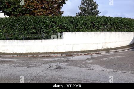 Zaun aus weißer Gipswand mit kleinem Geländer und Hecke oben, an der Straßenecke. Betonte Gehweg und Straße davor. Hintergrund Stockfoto