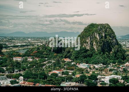 Da Nang, Vietnam - 14. August 2026. Ein dramatischer, von Grün bedeckter Kalksteingipfel erhebt sich über eine dichte tropische Stadtlandschaft. Stockfoto