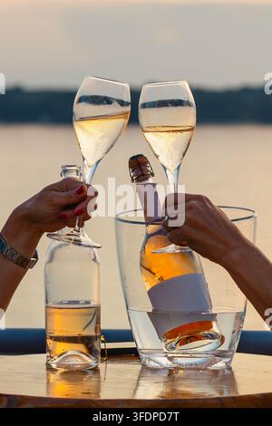 Zwei Personen stoßen mit Gläsern mit Sprudelwein oder Prosecco an während eines romantischen Moments am Meer. Eiskübel mit einer Flasche weißen Sprudelweins im Hintergrund Stockfoto