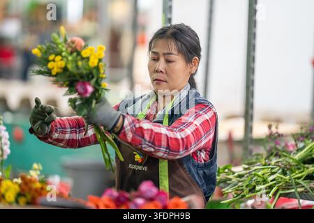 Blumenhändler arrangiert sorgfältig einen farbenfrohen gemischten Strauß, während er an einem geschäftigen Marktstand arbeitet, und trägt Schutzhandschuhe und eine Schürze. Die Szene c Stockfoto