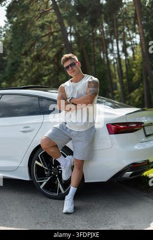 Attraktiver muskulöser Mann in Sonnenbrille, der mit verschränkten Armen an einem weißen Luxusauto auf einer sonnigen Waldstraße lehnt. Sportlicher Typ in Sportkleidung posiert draußen Stockfoto
