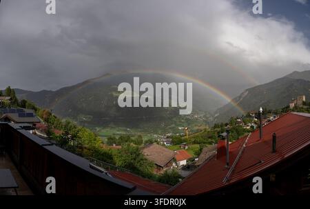 Ein lebhafter Doppelregenbogen wölbt sich über ein idyllisches Bergdorf und ein grünes Tal nach einem vorbeiziehenden Sturm und kontrastiert sonnenbeschienene Felder und Dächer mit düsterer Stockfoto