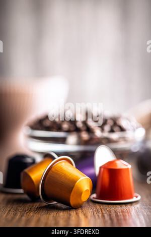 Bunte Kaffeekapseln und Espressopads, die auf einem Holztisch verstreut liegen, neben einer GlasSchale mit gerösteten Kaffeebohnen im weichen Fokus, die das morgendliche Ritual evozieren Stockfoto