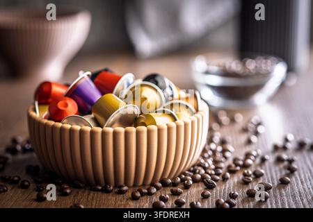Eine dekorative Schale gefüllt mit bunten Kaffeepads und Espressokapseln steht auf einem Holztisch mit verstreuten Kaffeebohnen und erinnert an Aroma, Morgen Stockfoto