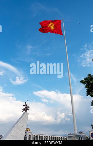 Niedrigwinkelaufnahme der nationalen Hauptflagge der Kirgisischen Republik, die im Wind auf einem riesigen Flaggenmast weht, mit dem Manas-Denkmal im Hintergrund. Stockfoto