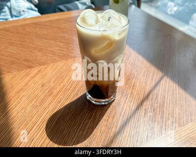 Hoher Eiskaffee in klarem Glas ruht auf sonnigem Holztisch, mit Eiswürfeln, geschichteten Kaffeetönen, warmen Reflexionen, weichem Café-Hintergrund und stark Stockfoto