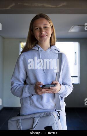 Teenage-Mädchen hält Telefon und schaut in Richtung Kamera im Flur, trägt grauen Hoodie und Messenger-Tasche Stockfoto