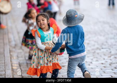 Zwei Kinder lachen und halten sich an den Händen, während sie in farbenfroher traditioneller peruanischer Kleidung auf einem Kopfsteinpflaster in Cusco tanzen, und erinnern an verspielte Kindheit Stockfoto