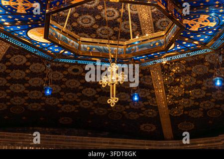 Das Innere der russisch-orthodoxen Kathedrale zeigt ein hängendes goldenes Kreuz unter einer gemusterten Kuppel mit blauem Sternenhimmel. Warmes Licht reflektiert auf vergoldeten Oberflächen Stockfoto