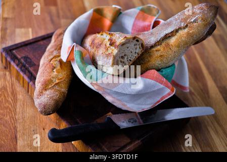 Sauerteig-Baguettes, die in einer mit einem Geschirrtuch ausgelegten Schüssel auf einem Holzschneidebrett arrangiert sind. Elegante Stillleben-Darstellung von frischem hausgemachtem Herdstein-Backgut Stockfoto