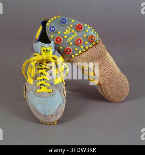 Ein Paar Sportschuhe, mit Stéphane Caristan, einem europäischen Athleten, der mit dem Bild verbunden ist, mit grauen und beigen Obermaterial, gelben Schnürsenkeln und einer Sohle mit bunten runden Nocken. Stockfoto